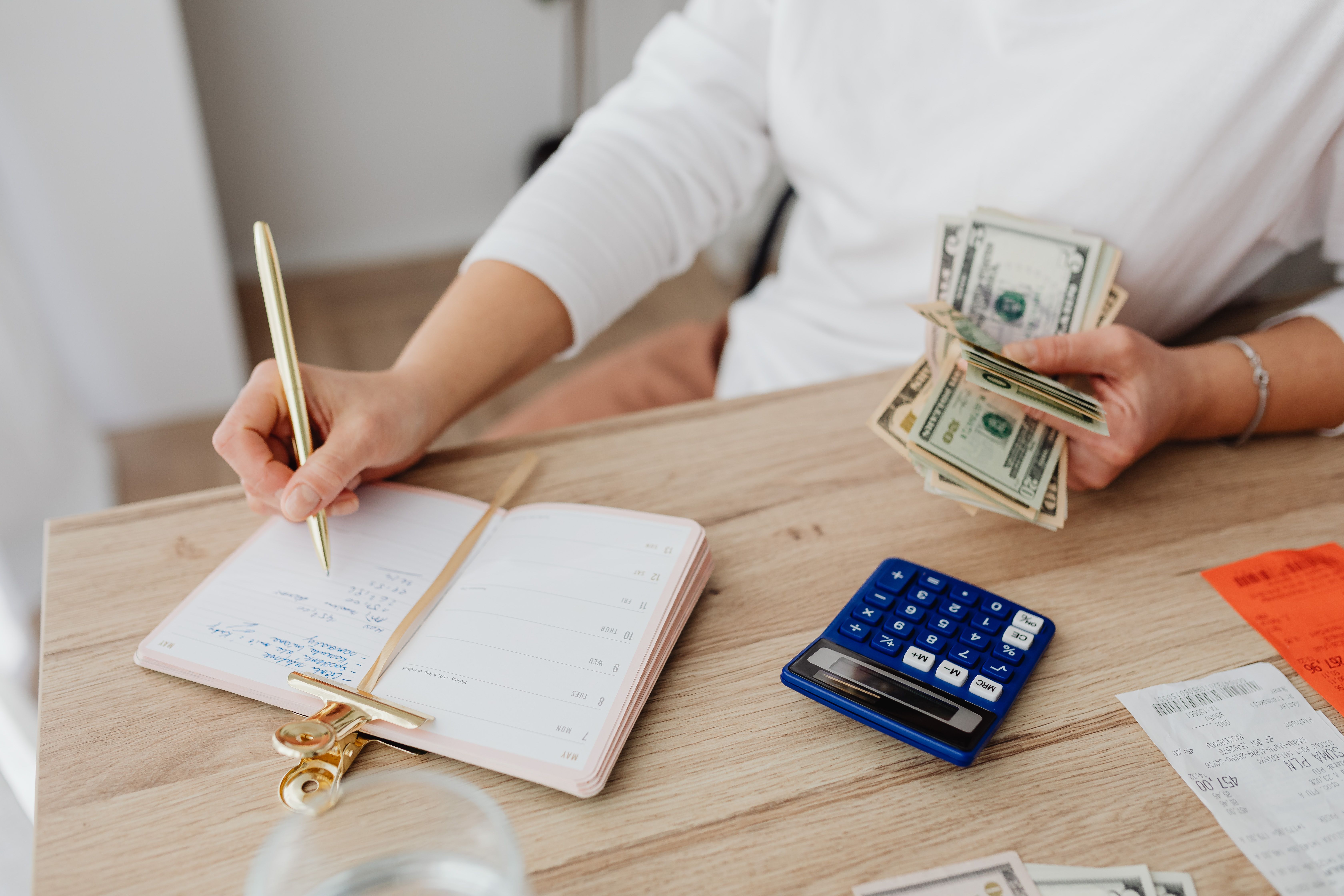 Person writing in a planner while using a calculator and holding cash at home